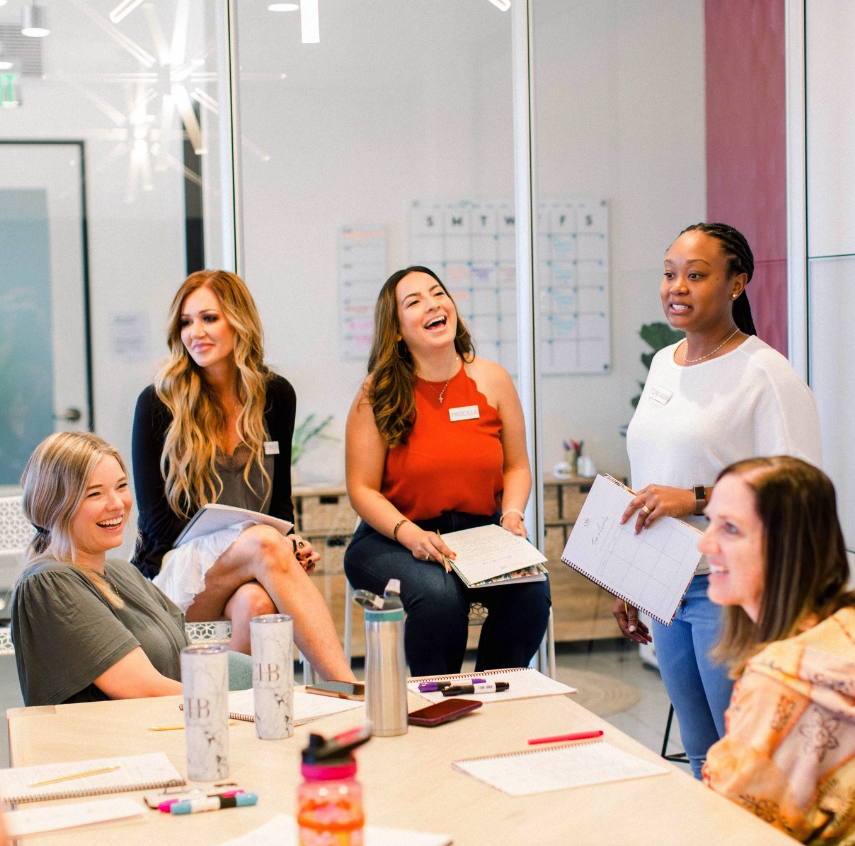 A group of women are gathered in a brightly lit co-working space. They are seated around a large table, engaged in lively conversation and laughing together. The atmosphere is casual and friendly, with some women holding coffee cups and others gesturing animatedly. The background shows modern office decor, including potted plants and shelves with books and office supplies. The women appear to be enjoying their time together, fostering a sense of community and collaboration.