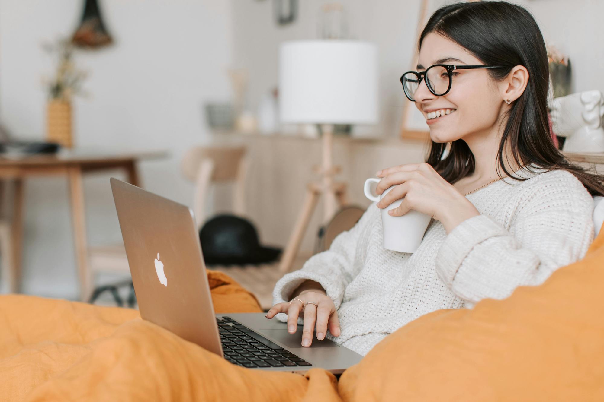 A young woman wearing glasses and a cozy white sweater sits comfortably with a warm yellow blanket while working on a laptop. She is smiling and holding a coffee cup in one hand, appearing relaxed and focused in a bright, modern home office setting. The background features minimalist furniture and soft lighting, creating a calm and inviting atmosphere for remote work.