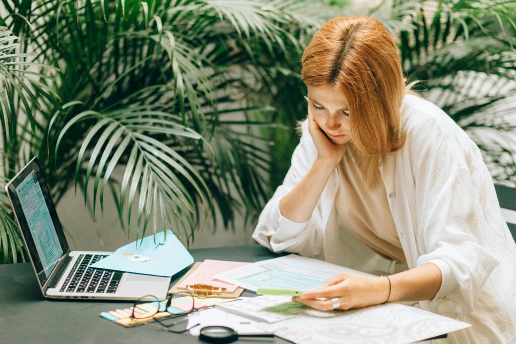 Woman sitting at a desk surrounded by papers, thoughtfully reviewing documents with a laptop open nearby, in a calm setting with lush green plants in the background.
This alt text highlights:
The subject (a woman)
The activity (thoughtfully reviewing papers)
The setting (desk, laptop, greenery)
The tone (calm, reflective—aligned with the blog post’s theme)