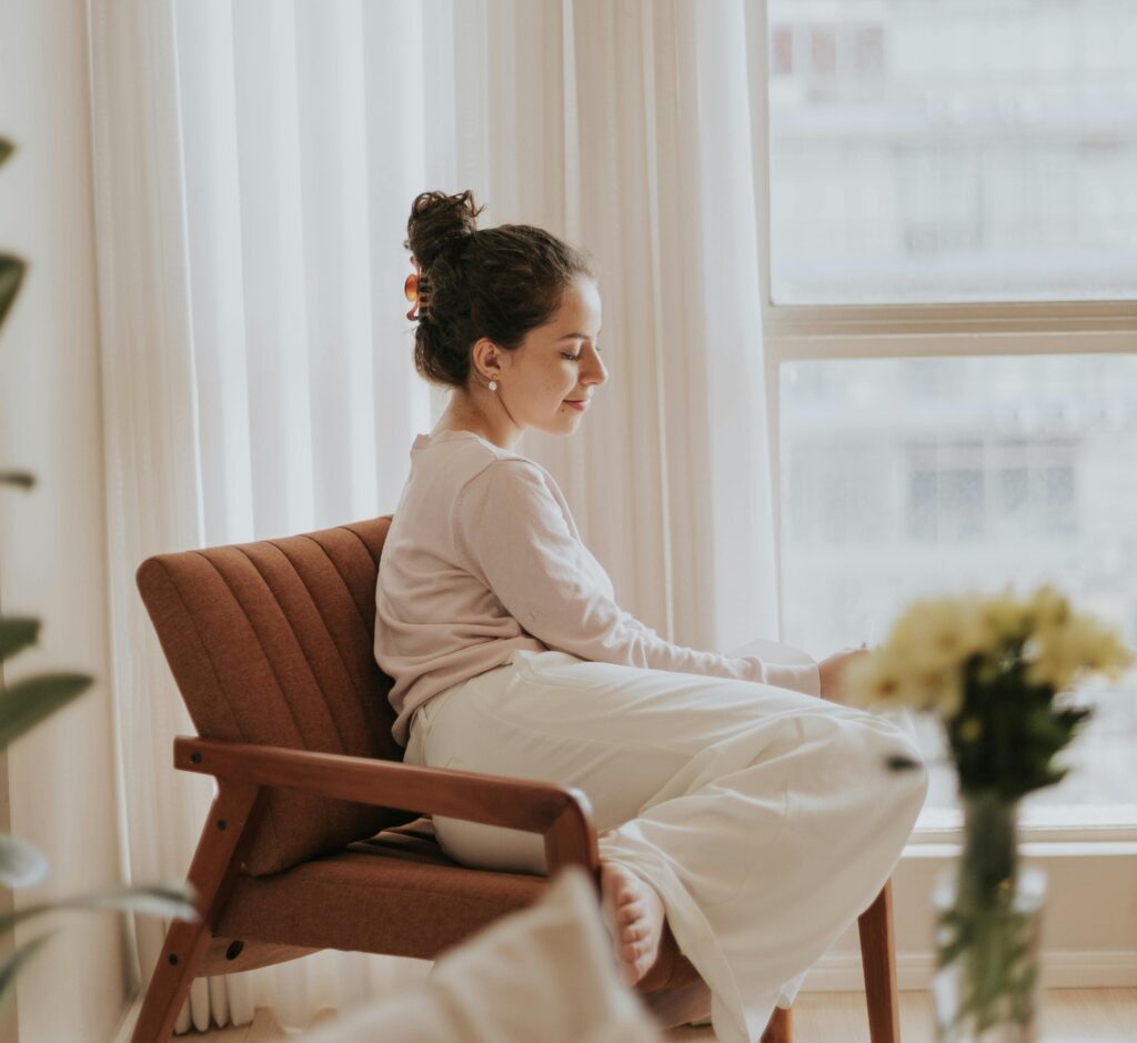Woman sitting quietly in a chair by a sunlit window, appearing calm and reflective.