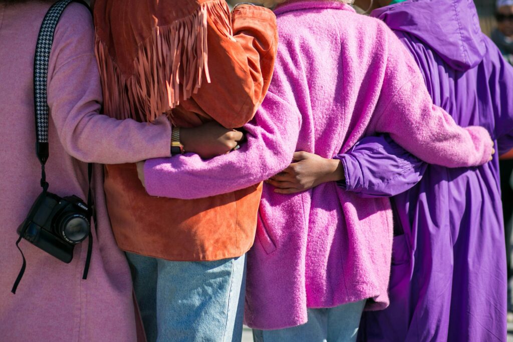 Women standing closely with arms linked, dressed in vibrant coats, reflecting warmth, belonging, and community support.