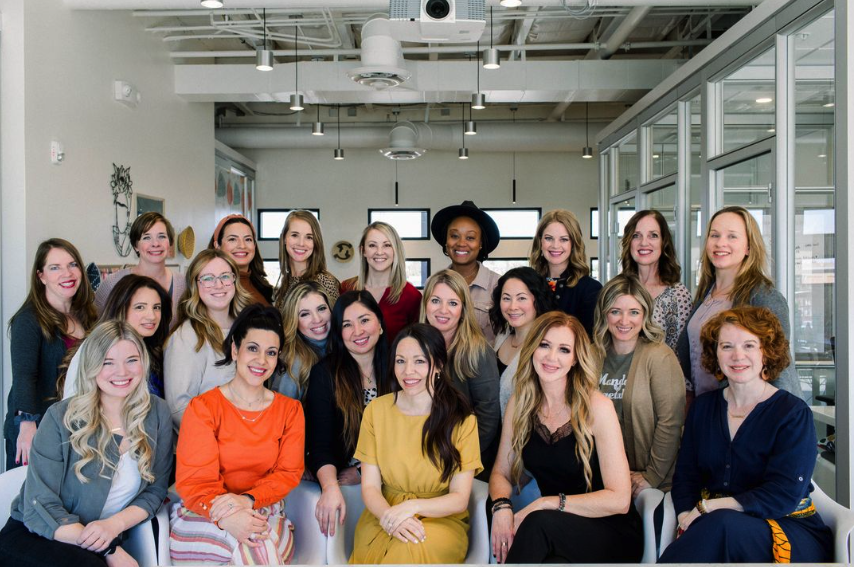 A group of women are posing for a group photo in a women's co-working space. They are standing close together, smiling warmly at the camera. The background features modern office decor, including large windows that let in plenty of natural light, potted plants, and bookshelves. The women are dressed in a variety of business casual attire, and some have their arms around each other, showing a sense of camaraderie and connection. The overall mood is positive and empowering.