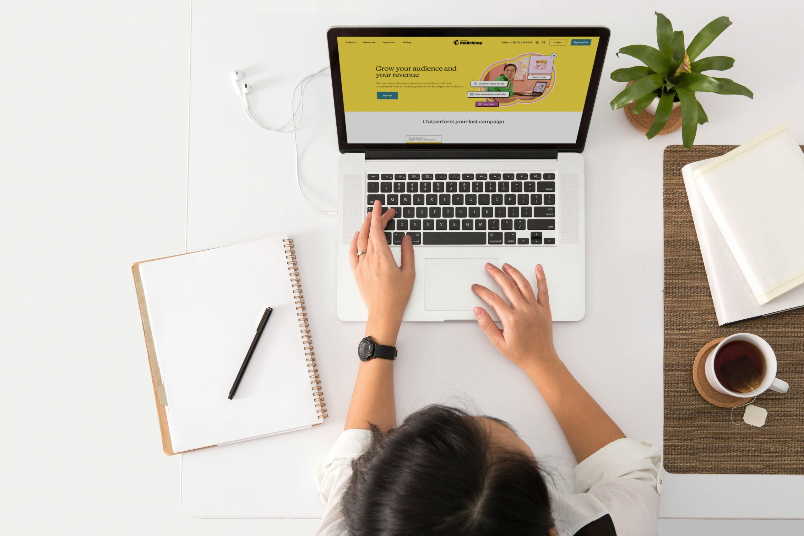 Overhead view of a woman working at a modern, minimalistic desk setup with a laptop displaying the Mailchimp website. She has a notebook, a pen, a cup of tea, and a small plant nearby, creating a clean and productive workspace.